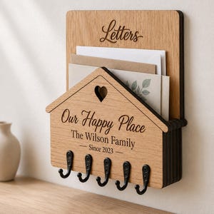 Puede incluir: Un organizador de correo y llavero de pared de madera clara con cinco ganchos de metal negro. El panel frontal tiene forma de casa con un recorte de coraz&oacute;n y el texto grabado "Our Happy Place", "The Wilson Family" y "Since 2023". El panel superior tiene grabado "Letters".