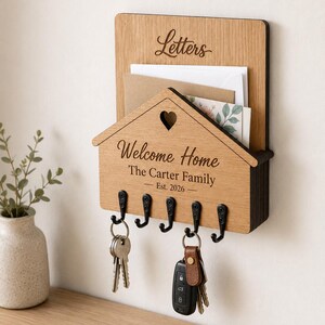 Puede incluir: Un organizador de correo y llavero de pared de madera marr&oacute;n claro. La secci&oacute;n superior tiene la palabra Letters. La secci&oacute;n inferior con forma de casa presenta un recorte de coraz&oacute;n, el texto Welcome Home, The Carter Family y Est. 2026.
