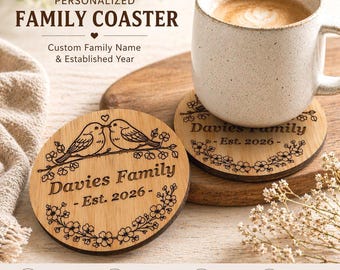 Puede incluir: Posavasos familiar personalizado de madera con grabado láser. Presenta un diseño floral con dos pájaros y el texto "Davies Family - Est. 2026." Una taza blanca con café descansa sobre el posavasos. El texto "Personalized Family Coaster" es visible.