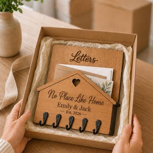 Puede incluir: Un organizador de pared de madera en una caja de cart&oacute;n. La pieza trasera est&aacute; grabada con la palabra Letters. La pieza frontal en forma de casa con un recorte de coraz&oacute;n est&aacute; grabada con No Place Like Home, Emily & Jack, Est. 2026 y cinco ganchos de metal negro.