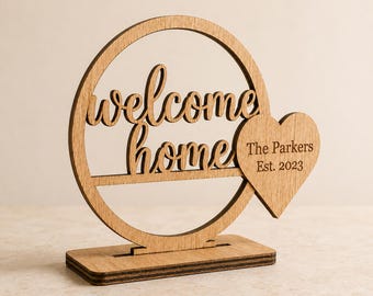 Puede incluir: Un letrero de mesa de madera de color marrón claro con un marco circular con el texto recortado "welcome home" y una pieza en forma de corazón a la derecha grabada con el texto "The Parkers Est. 2023", montado sobre una base rectangular.