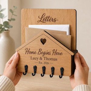 Puede incluir: Un organizador de pared de madera de color marr&oacute;n claro con un panel frontal en forma de casa y un recorte de coraz&oacute;n. Est&aacute; grabado con el texto "Letters", "Home Begins Here", "Lucy & Thomas" y "Est. 2026". Cinco ganchos de metal negro est&aacute;n fijados en la parte inferior.