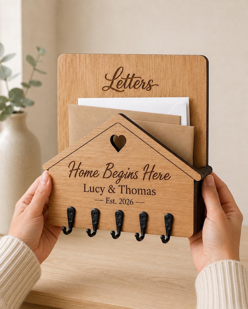 Puede incluir: Un organizador de pared de madera de color marr&oacute;n claro con un panel frontal en forma de casa y un recorte de coraz&oacute;n. Est&aacute; grabado con el texto "Letters", "Home Begins Here", "Lucy & Thomas" y "Est. 2026". Cinco ganchos de metal negro est&aacute;n fijados en la parte inferior.