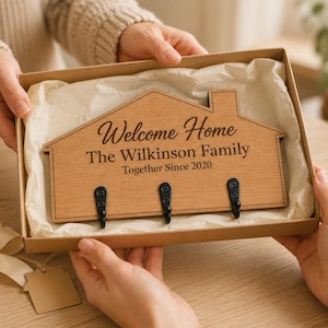 Puede incluir: Un perchero de pared de madera con forma de casa y tres ganchos de metal negro. La madera está grabada con el texto "Welcome Home", "The Wilkinson Family" y "Together Since 2020". El artículo se presenta dentro de una caja de regalo de cartón con papel de seda.