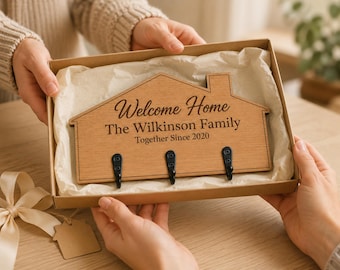 Puede incluir: Un perchero de pared de madera con forma de casa y tres ganchos de metal negro. La madera está grabada con el texto "Welcome Home", "The Wilkinson Family" y "Together Since 2020". El artículo se presenta dentro de una caja de regalo de cartón con papel de seda.