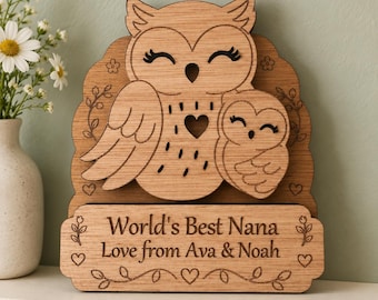 Puede incluir: Una placa de madera de color marrón claro con un diseño de búho y cría cortado con láser, adornada con motivos florales y corazones grabados. La base tiene grabada la inscripción: World's Best Nana, Love from Ava & Noah, rodeada de motivos.