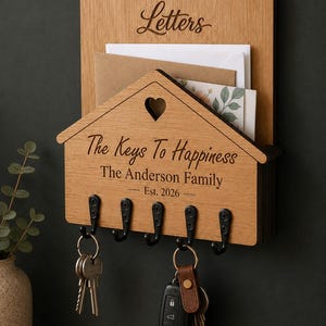 Puede incluir: Un organizador de correo y llavero de pared de madera marr&oacute;n claro. La secci&oacute;n superior est&aacute; grabada con la palabra Letters. La secci&oacute;n inferior en forma de casa tiene un recorte de coraz&oacute;n, el texto The Keys To Happiness, The Anderson Family, Est. 2026 y cinco ganchos de metal negro con dos juegos de llaves.