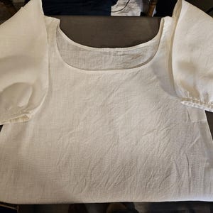 May include: A white blouse featuring a wide scoop neckline and voluminous puff sleeves. The garment is crafted from a textured fabric with visible weave details, showcasing a simple, clean design with gathered cuffs at the ends of the sleeves.
