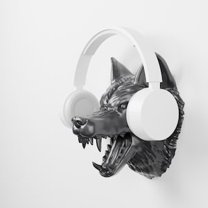 May include: A black wolf head wall mount sculpture with an open mouth showing teeth, wearing a pair of solid white over-ear headphones. The wolf head is detailed with textured fur and pointed ears, positioned against a plain white background.