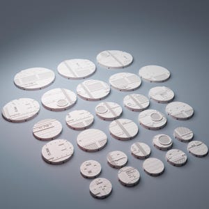 May include: A collection of twenty-four circular white plastic bases for miniature figures, each featuring distinct moulded textures including grid patterns, vents, panels, and industrial surface details in various sizes arranged on a flat surface.