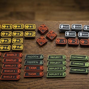 May include: A selection of game tokens with different labels and symbols. The tokens are rectangular and diamond-shaped, with text such as "BUFF", "STUNNED", and numbers. Colours include yellow, black, red, and green. The tokens are arranged on a wooden surface.
