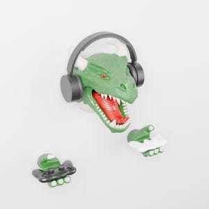 May include: A green dinosaur head wall mount wearing black over-ear headphones, featuring a wide-open mouth with red interior and white teeth. Two separate green dinosaur claw hands are positioned below, one holding a black game controller.