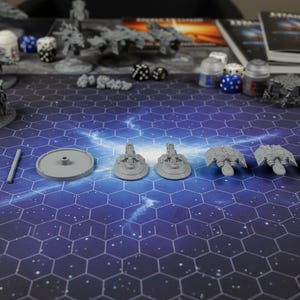 May include: A tabletop gaming scene featuring miniature spaceship models, dice, and terrain pieces. The scene is set on a hexagonal grid mat with a space-themed design. Various gray plastic models are arranged for gameplay.