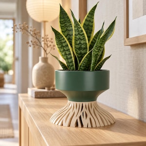 May include: A snake plant with variegated green and yellow leaves potted in a dark green cylindrical planter. The planter features a wide, light-colored base with a decorative, intricate cutout pattern resembling organic root structures.