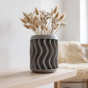 May include: A matte dark gray cylindrical vase with a vertical wavy, ribbed texture on the exterior. The vase is filled with a bouquet of dried beige bunny tail grass and small white flowers, creating a neutral and textured arrangement.