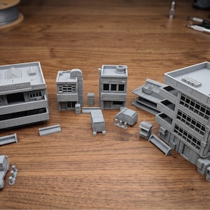 May include: A selection of grey miniature buildings, including a multi-storey structure, several smaller buildings, and a few vehicles. The models are arranged on a wooden surface, suggesting a cityscape or architectural model.