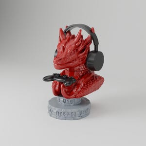 May include: A red dragon bust figurine wearing black over-ear headphones and holding a black game controller. The dragon is mounted on a circular gray stone-textured base with carved runic symbols around the perimeter of the stand.