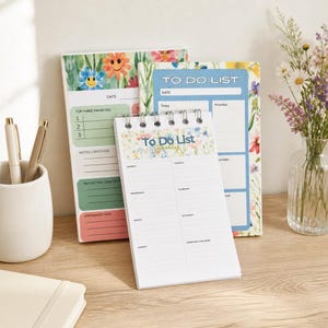 May include: A collection of floral-themed to-do list notepads. One notepad is spiral-bound, displaying a weekly layout. Two other notepads feature a daily layout with sections for priorities and notes. Pens and a vase of flowers are also present.