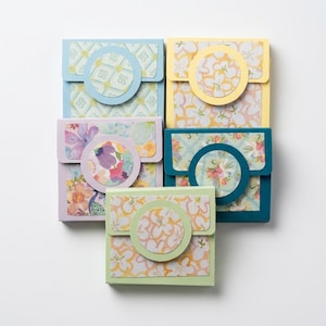May include: Five square paper envelopes with flap closures, each featuring a circular accent piece. The envelopes display various patterns including floral, geometric, and abstract designs in shades of blue, yellow, teal, lavender, and light green.