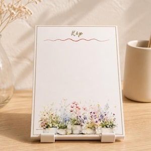 May include: A white notepad with a floral design at the bottom and the word "Kip" at the top. The floral design features a variety of colorful flowers in small white pots. The notepad is propped up by a white stand.