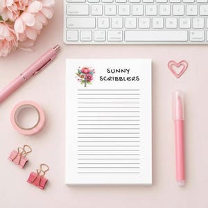 May include: A white notepad with lined pages and the text "SUNNY SCRIBBLERS" in black. The notepad is decorated with a floral design. A pink pen, tape, paper clips, and a marker are arranged around the notepad on a pink surface.