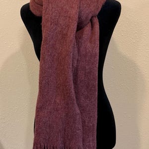 May include: A burgundy scarf with fringe detail, draped over a black mannequin. The scarf is made of a soft, textured material. The scarf is a solid color and appears to be warm and cozy.