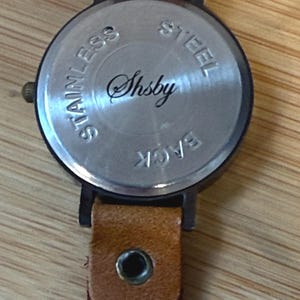 May include: Close-up of the back of a wristwatch with a brown leather band. The watch face is silver with the word "Shsby" in cursive. The words "Stainless Steel Back" are also visible.