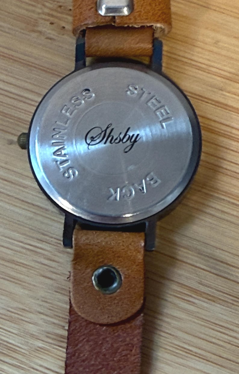 May include: Close-up of the back of a wristwatch with a brown leather band. The watch face is silver with the word "Shsby" in cursive. The words "Stainless Steel Back" are also visible.