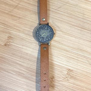 May include: A brown leather strap watch with a round, dark-colored face. The watch face features numbers and hands, and the strap has a buckle and several adjustment holes. The watch is displayed on a wooden surface.