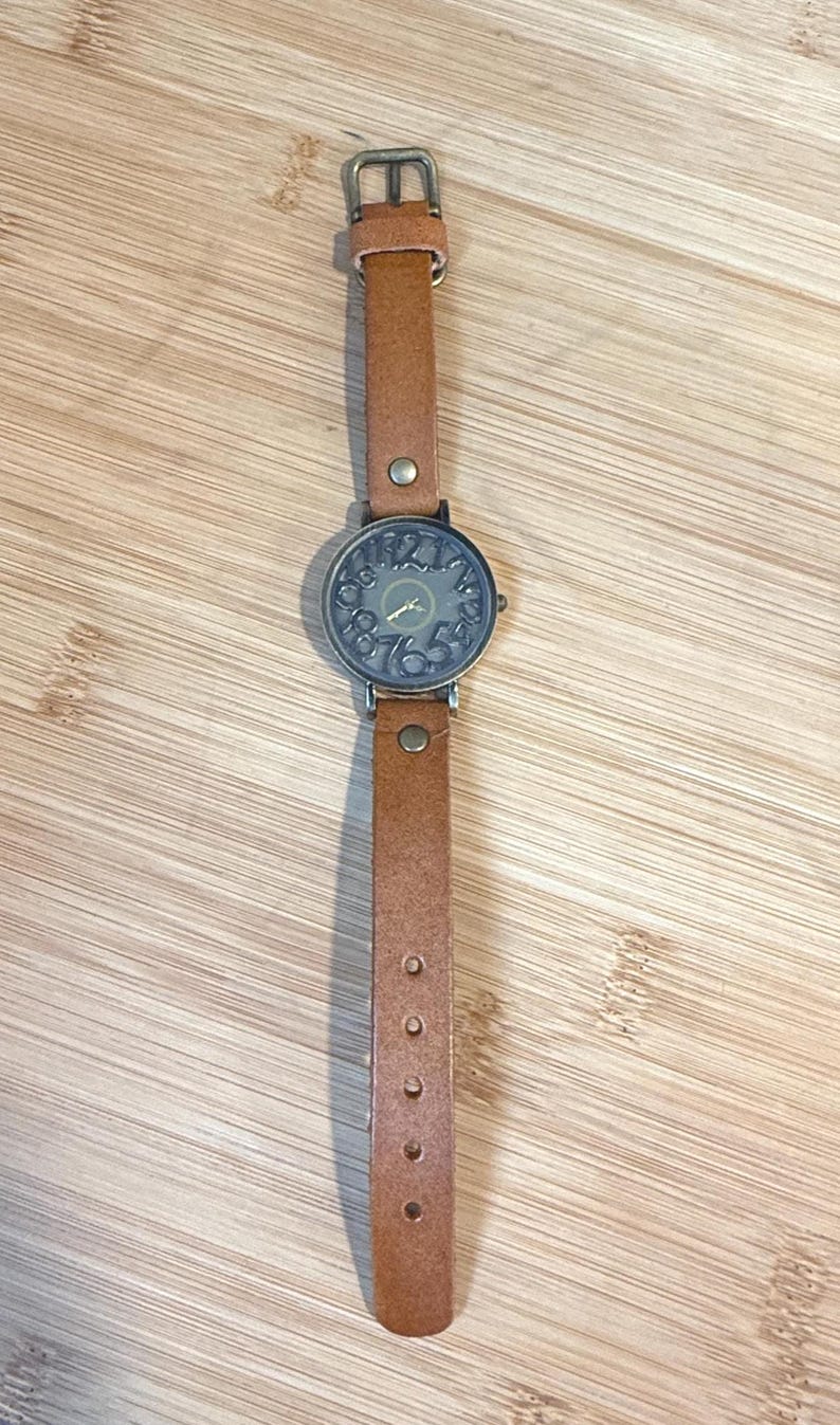 May include: A brown leather strap watch with a round, dark-colored face. The watch face features numbers and hands, and the strap has a buckle and several adjustment holes. The watch is displayed on a wooden surface.