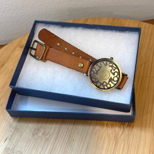 May include: A brown leather watch with a brass-colored face and numbers, presented in a navy blue box with white padding. The watch band is a light brown color with a brass buckle.