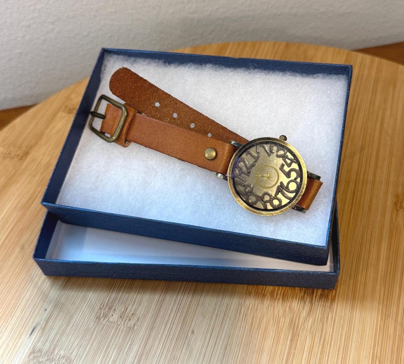 May include: A brown leather watch with a brass-colored face and numbers, presented in a navy blue box with white padding. The watch band is a light brown color with a brass buckle.