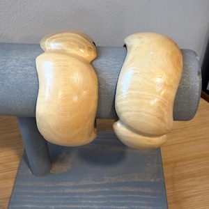 May include: Two light-colored, polished wooden bangle bracelets with a smooth, curved, and organic shape. The bracelets feature a natural wood grain pattern with variations in tone, showcasing a rounded, thick profile that encircles the wrist area.