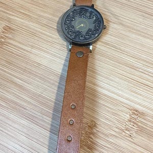 May include: A vintage-style wristwatch with a brown leather band. The watch face is a dark bronze color with ornate numerals and hands. The band has a buckle closure and measures approximately 9 inches in length.