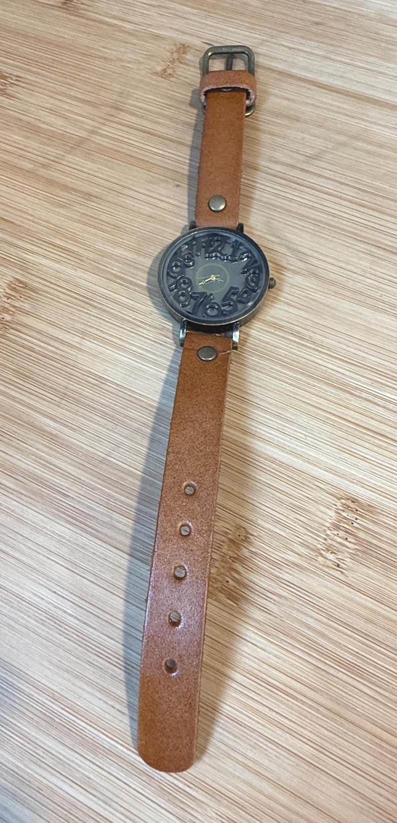 May include: A vintage-style wristwatch with a brown leather band. The watch face is a dark bronze color with ornate numerals and hands. The band has a buckle closure and measures approximately 9 inches in length.
