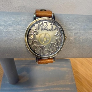 May include: A vintage-style wristwatch with a brown leather band. The watch face has a bronze-colored frame with large, open-style numerals. The watch is displayed on a gray wooden stand.
