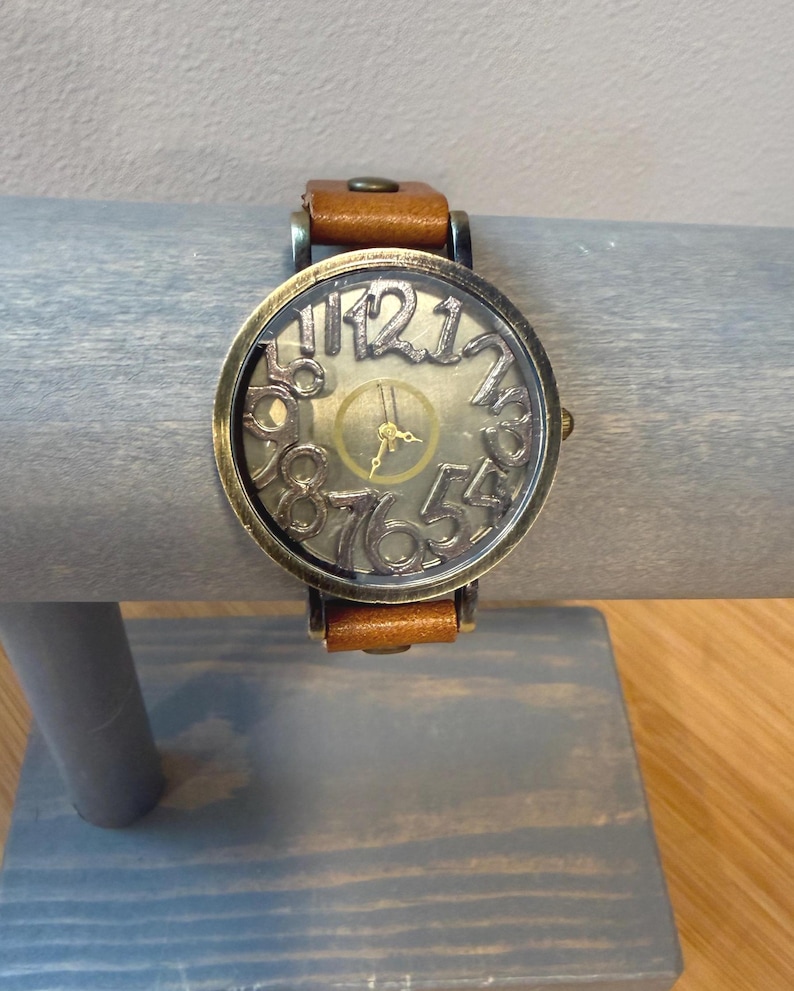 May include: A vintage-style wristwatch with a brown leather band. The watch face has a bronze-colored frame with large, open-style numerals. The watch is displayed on a gray wooden stand.