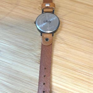 May include: A wristwatch with a brown leather band and a round, silver-toned watch face. The watch band has a buckle closure and multiple adjustment holes. The watch face has the word "Shsby" engraved on it.