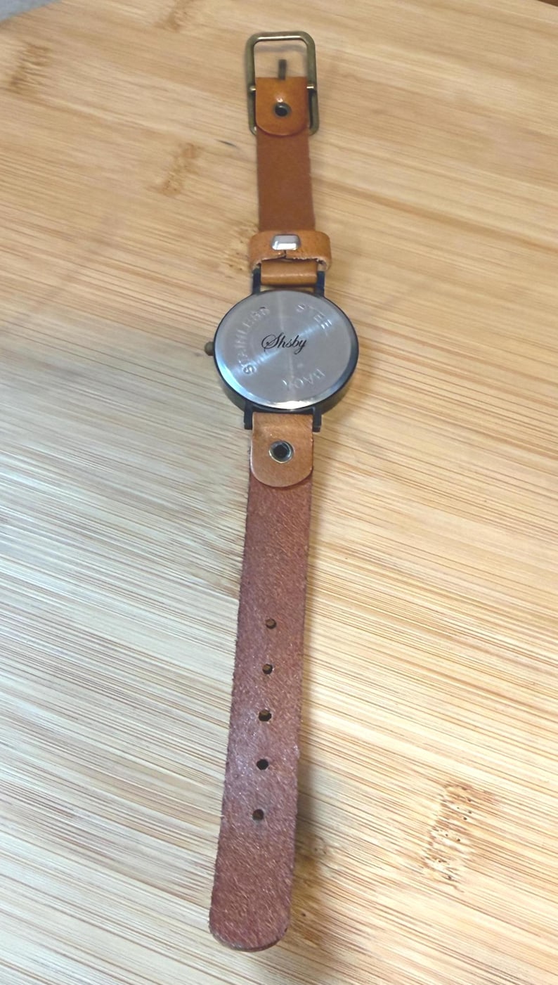 May include: A wristwatch with a brown leather band and a round, silver-toned watch face. The watch band has a buckle closure and multiple adjustment holes. The watch face has the word "Shsby" engraved on it.