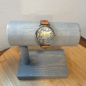 May include: A vintage-style wristwatch with a bronze-colored face and a brown leather band. The watch face features large, stylized numerals. The watch is displayed on a gray wooden watch stand.