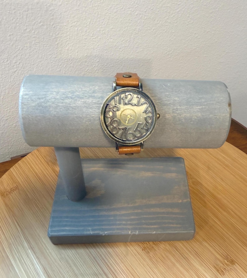 May include: A vintage-style wristwatch with a bronze-colored face and a brown leather band. The watch face features large, stylized numerals. The watch is displayed on a gray wooden watch stand.