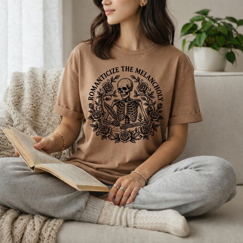 Puede incluir: Camiseta color beige con un dise&ntilde;o negro de un esqueleto sosteniendo una pluma, rodeado de rosas. El texto "ROMANTICIZE THE MELANCHOLY" est&aacute; sobre el esqueleto. La persona lleva la camiseta y est&aacute; sentada en un sof&aacute;.