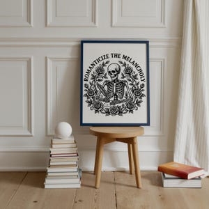Puede incluir: Una impresi&oacute;n art&iacute;stica enmarcada con un marco azul oscuro. La obra de arte en blanco y negro presenta un esqueleto escribiendo con una pluma, rodeado de rosas, con el texto "ROMANTICIZE THE MELANCHOLY". La impresi&oacute;n se exhibe en un taburete de madera.