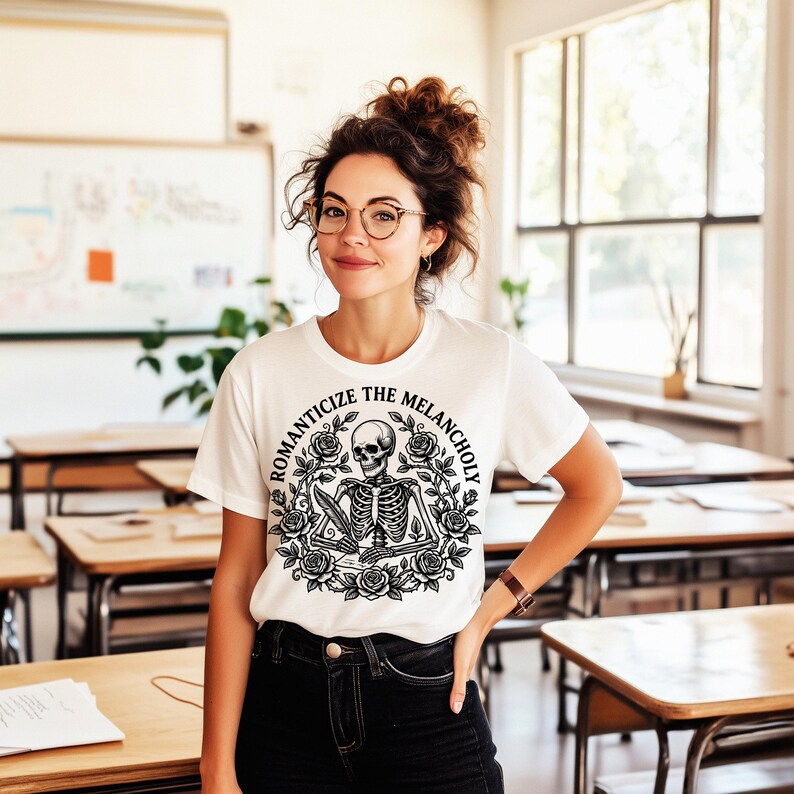 Puede incluir: Camiseta blanca con un dise&ntilde;o negro de un esqueleto con una pluma, rodeado de rosas y el texto "ROMANTICIZE THE MELANCHOLY". La camiseta es de cuello redondo.