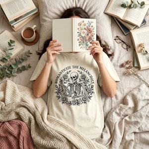 Puede incluir: Una camiseta color crema con un dise&ntilde;o de esqueleto en blanco y negro rodeado de rosas, con el texto "ROMANTICIZE THE MELANCHOLY". La imagen tambi&eacute;n incluye un libro con ilustraciones florales, libros y una taza de caf&eacute;.