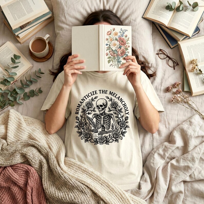 Puede incluir: Una camiseta color crema con un dise&ntilde;o de esqueleto en blanco y negro rodeado de rosas, con el texto "ROMANTICIZE THE MELANCHOLY". La imagen tambi&eacute;n incluye un libro con ilustraciones florales, libros y una taza de caf&eacute;.