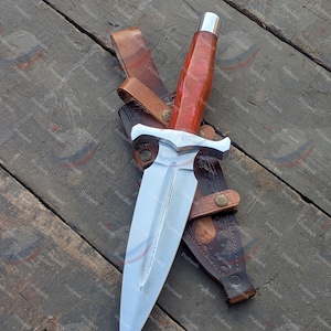 May include: A dagger with a polished steel blade and a red and silver handle, resting in a brown leather sheath. The sheath has a belt loop and decorative stitching. The dagger is displayed on a wooden surface.
