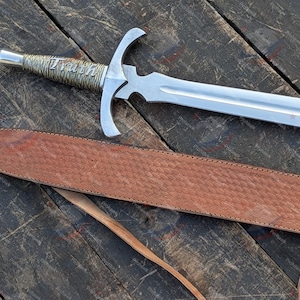 Può includere: Una spada in metallo color argento con guardia a croce e impugnatura testurizzata con la parola Truth incisa in corsivo. La spada è accompagnata da un lungo fodero in pelle marrone con un motivo in rilievo ripetuto e sottili cinghie.