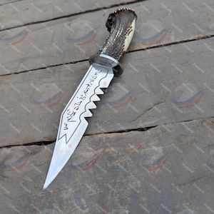 Peut inclure: Un couteau de chasse artisanal avec un manche en corne texturé et une lame en acier inoxydable. La lame présente un bord dentelé et du texte gravé. Le couteau est présenté sur une surface en bois.
