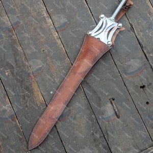 May include: A medieval-style sword with a silver blade and handle, in a brown leather sheath. The sword's hilt has a decorative design. The sheath is stitched with white thread. The sword rests on a weathered wooden surface.